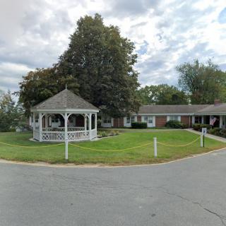 A scenic view of a gazebo on a lawn with a building in the background under a cloudy sky.