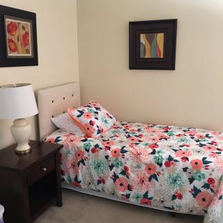 A cozy bedroom with a single bed featuring floral bedding, a nightstand with a lamp, and decorative wall art.