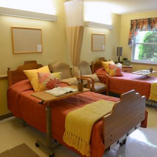 A well-lit hospital room with two beds, each equipped with pillows and blankets, separated by a privacy curtain. The room includes chairs and a window providing natural light.