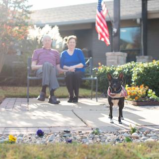 A couple sitting on a patio with their dog and an American flag displayed in the garden.
