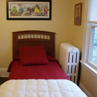 A cozy bedroom featuring a single bed with red bedding, a radiator by the window, and wall art.