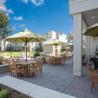 An outdoor patio area with tables, chairs, and umbrellas in front of a building on a sunny day.