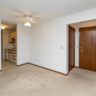 A simple apartment interior featuring a living room with carpet flooring, white walls, a ceiling fan, and a view of the kitchen and entrance door.