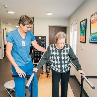 An elderly person receives assistance from a healthcare professional during a physical therapy session indoors.