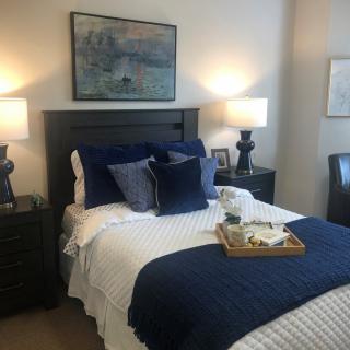 A cozy and well-decorated bedroom featuring a neatly made bed with blue and white bedding, accented by two bedside lamps and artwork above the headboard.