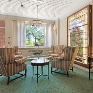 A cozy living room with armchairs around a small table, featuring a large stained glass window and abundant natural light.