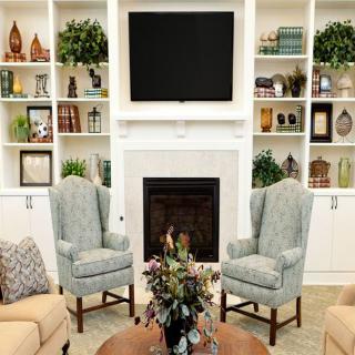A cozy living room featuring a central fireplace, two armchairs, and a TV mounted above the mantel. The built-in shelves are filled with various books and decorative items.