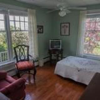 A cozy bedroom with large windows, featuring a bed, chairs, and a television on hardwood flooring.