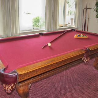 A pool table with red felt, featuring a cue stick and triangle rack of balls in a well-lit room.