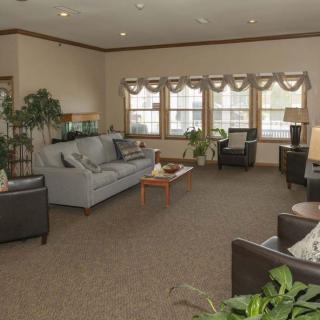 A cozy living room with sofas, chairs, and a coffee table surrounded by large windows and indoor plants.