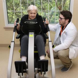 An elderly person receiving guidance from a healthcare professional while using exercise equipment indoors.