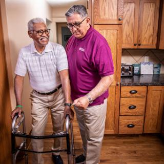 A caregiver assists an elderly man using a walker in a warm, inviting home.