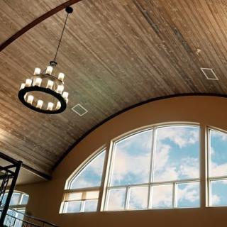 A spacious room with a wooden ceiling and an elegant chandelier, featuring large arched windows letting in natural light.