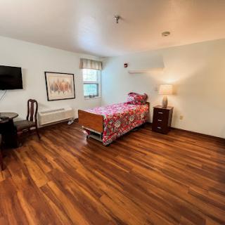 A spacious and well-furnished bedroom featuring a wooden floor, a bed with colorful bedding, a TV mounted on the wall, and a bedside lamp.