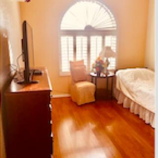 A cozy bedroom with hardwood floors, featuring a bed, armchair, television, and large window allowing natural light to filter through.