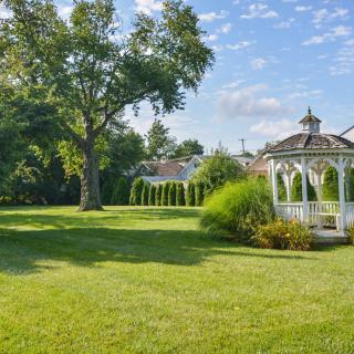 A serene garden scene featuring a white gazebo, lush trees, and a well-maintained lawn under a clear blue sky.