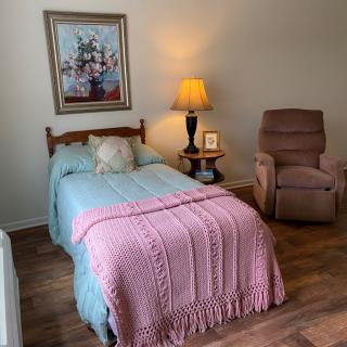 A cozy bedroom featuring a bed with a pink blanket, a recliner chair, a lamp on a side table, and artwork on the wall.