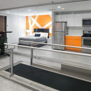 Modern apartment interior with a compact kitchen and bedroom area, featuring stainless steel appliances and bright orange accents.