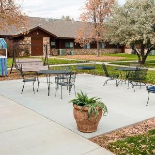 An outdoor patio area with metal tables and chairs, surrounded by trees and a building in the background.