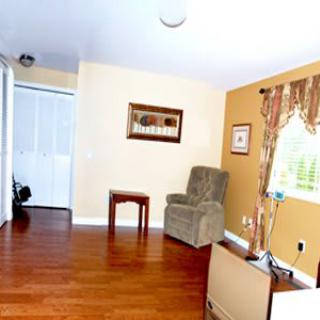 A cozy living room with hardwood floors, featuring an armchair, side table, and sunlight streaming through a window with decorative curtains.