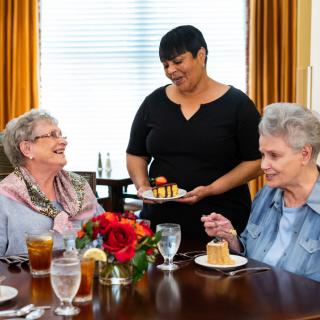 Two elderly women enjoy a meal and laughter as a waitress serves dessert at a restaurant.