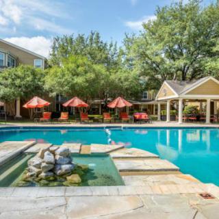 A serene outdoor swimming pool area at an apartment complex with patio seating and lush greenery.