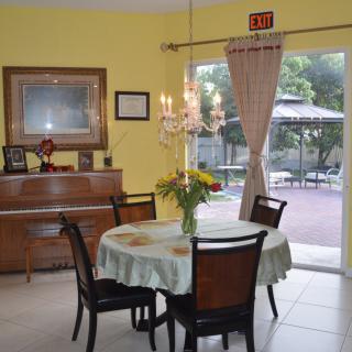 A cozy dining room with a round table, chairs, and a piano against yellow walls. A chandelier hangs overhead, and curtains open to reveal a view of the patio.