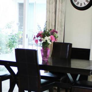 A dining room featuring a dark wooden table with chairs, adorned with a vase of vibrant flowers and a wall clock in the background.