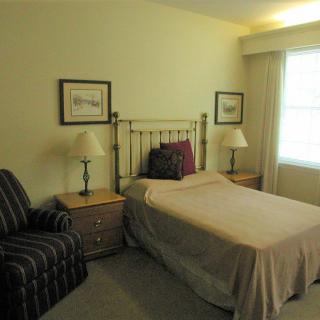 A cozy bedroom featuring a single bed with a metal frame, flanked by two nightstands and lamps. A comfortable chair is positioned next to the bed, and light pours in through a window.