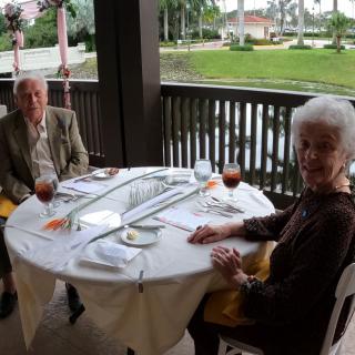 An elderly couple enjoys a meal at an outdoor waterfront restaurant.