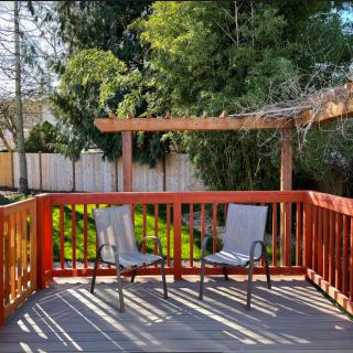 A sunny outdoor deck with two chairs and a pergola, overlooking a garden.