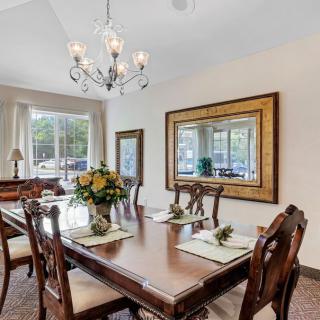 A well-lit dining room featuring a large wooden table with elegant chairs, a chandelier, and decorative elements such as mirrors and flowers.