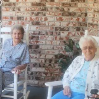 Two elderly women sitting in rocking chairs on a porch with a brick wall background.