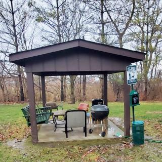 A small outdoor gazebo with chairs and a grill set in a park surrounded by trees.