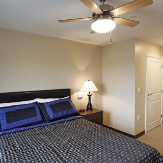 A cozy bedroom with a neatly made bed, bedside lamp, and ceiling fan.