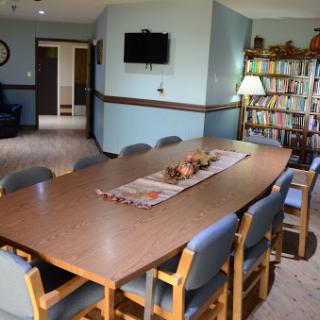 A spacious dining room featuring a large wooden table surrounded by chairs, with a bookshelf and television in the background.