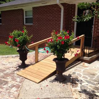 A wooden accessibility ramp leading into a brick house, flanked by two large planters with vibrant red flowers.