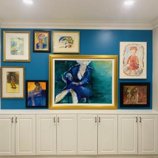 A well-arranged collection of paintings displayed on a blue accent wall above white cabinets, showcasing an eclectic mix of art styles.