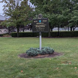 A historical marker stands in a green park, surrounded by trees.