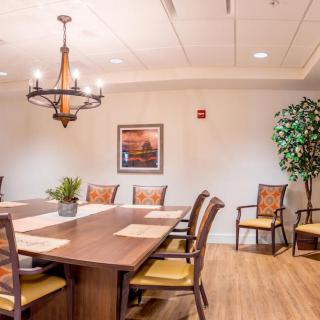 A well-furnished conference room with a large wooden table, cushioned chairs, a chandelier, and decorative plants.