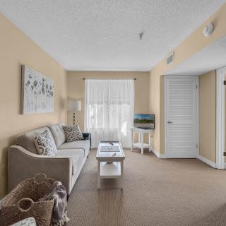 A cozy living room with beige walls, featuring a sofa and coffee table, decorated with wall art and illuminated by natural light through window blinds.