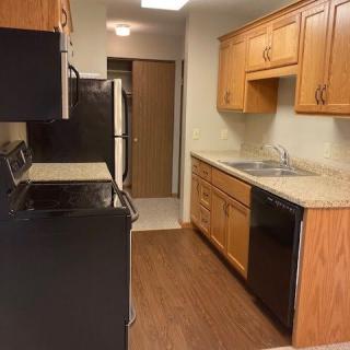 A narrow kitchen with wood cabinets and black appliances, featuring a stove, refrigerator, sink, and dishwasher.