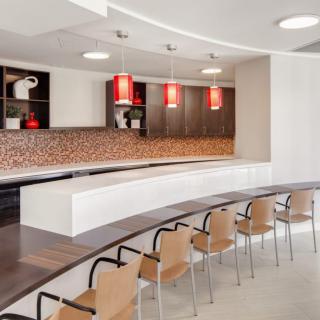 A modern kitchen interior with a long countertop, pendant lights, and rows of chairs.