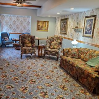 A cozy living room with floral patterned furniture and carpet, featuring armchairs, a sofa, decorative wallpaper, paintings, and a ceiling fan.