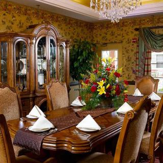 An elegant dining room with a large wooden table, ornate chairs, and a chandelier overhead. A floral arrangement serves as the centerpiece on the table.