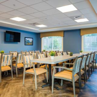 A spacious dining room with arranged tables and chairs, featuring a television on the wall and large windows.