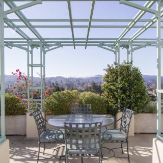 A picturesque outdoor patio with a pergola, set with a table and chairs, surrounded by greenery and offering a scenic view.