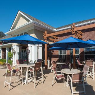 An outdoor patio area with tables and chairs shaded by blue umbrellas, set against the backdrop of a building on a sunny day.