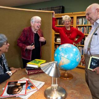A group of seniors engaged in a lively discussion around a table with books and a globe in a library setting.