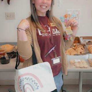 A cheerful woman poses with a bag in front of a table filled with baked goods.